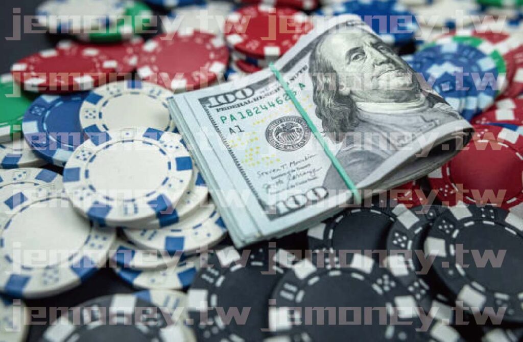 「畫面中央為一疊綁橡皮筋的百元美鈔，周圍散落紅藍白黑四色籌碼。」